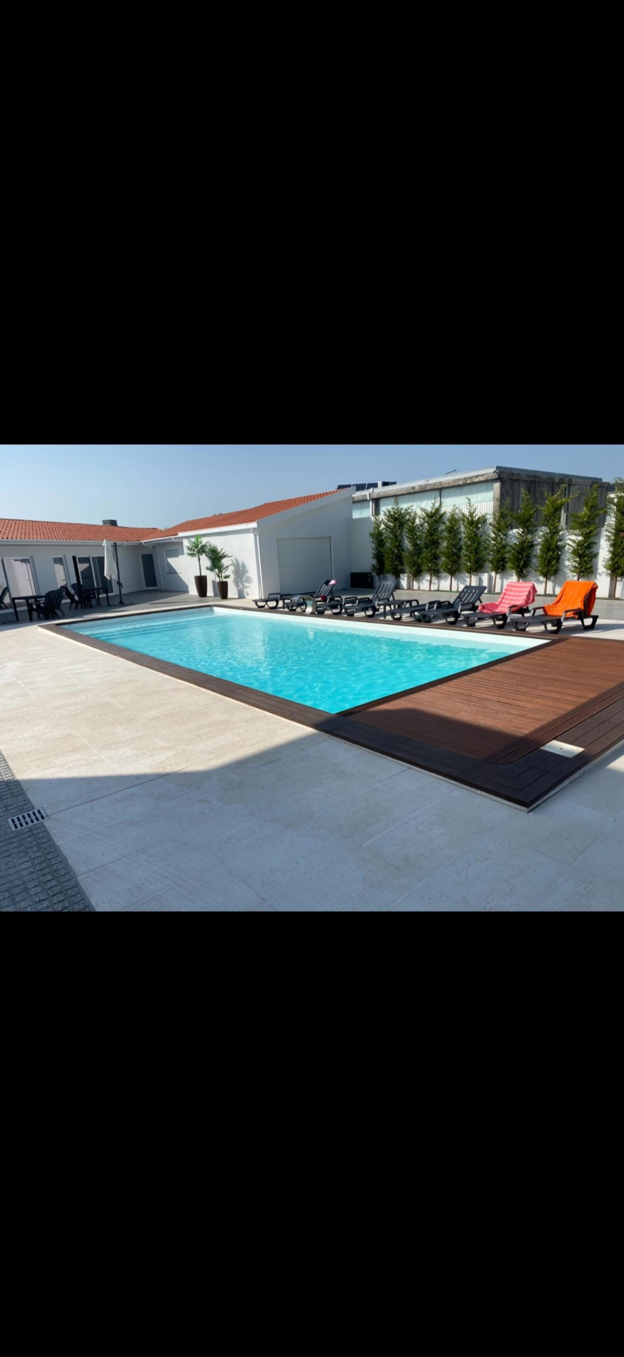 Piscina retangular azul com bordas de madeira, espreguiçadeiras coloridas e plantas em pátio ensolarado.